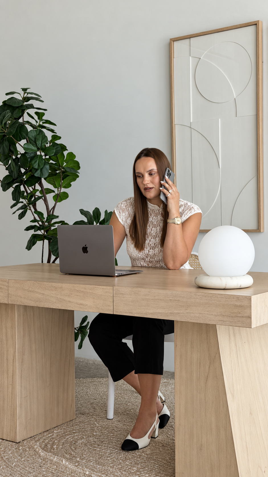 A woman sits at a light wood desk in a bright home office, talking on a smartphone with a laptop in front of her. She wears a textured cream top and black cap-toe heels.